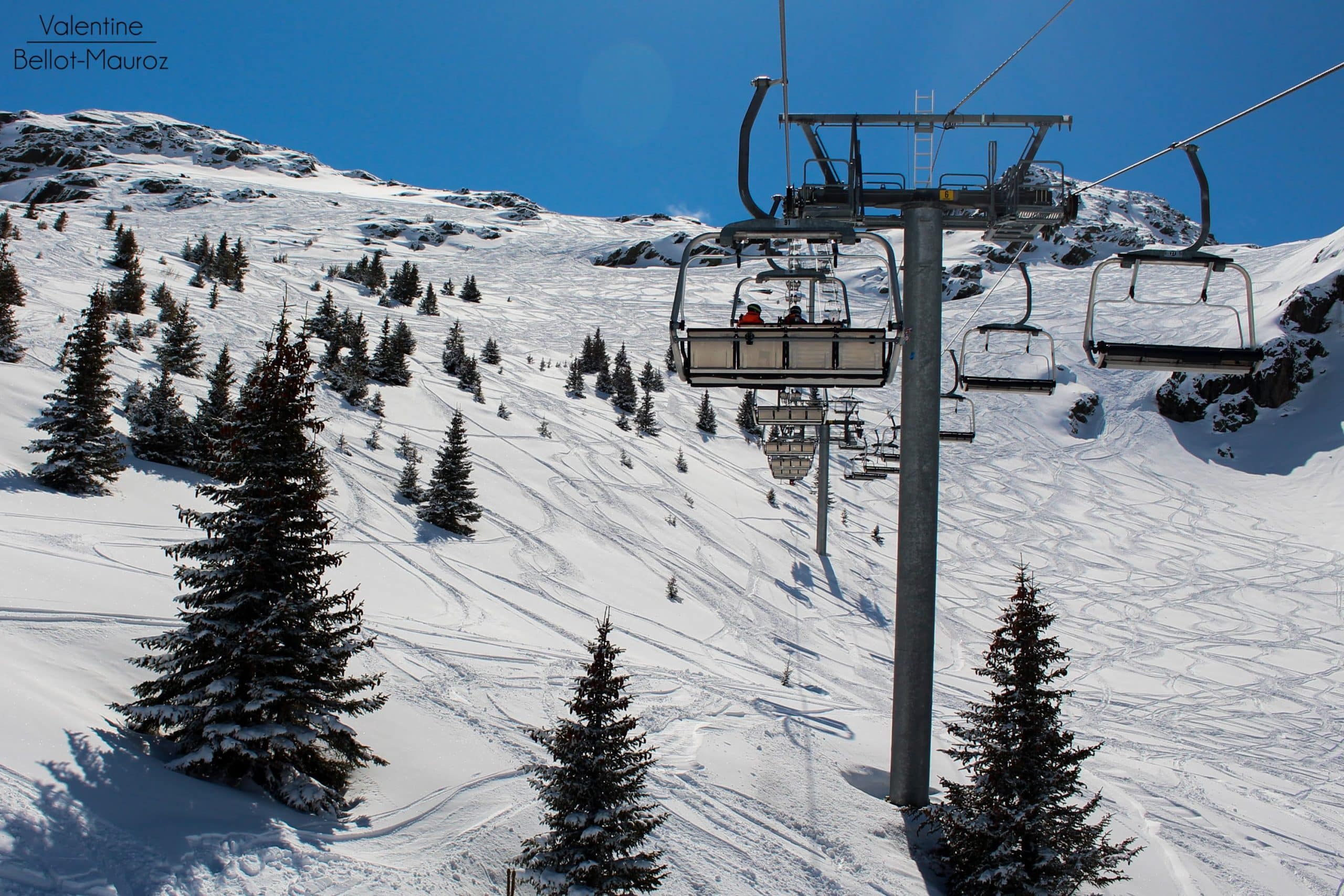 Valentine Bellot-Mauroz. Des passagers montent en télésiège sur une pente de ski enneigée, parsemée de sapins et de nombreuses traces, sous un ciel bleu éclatant.