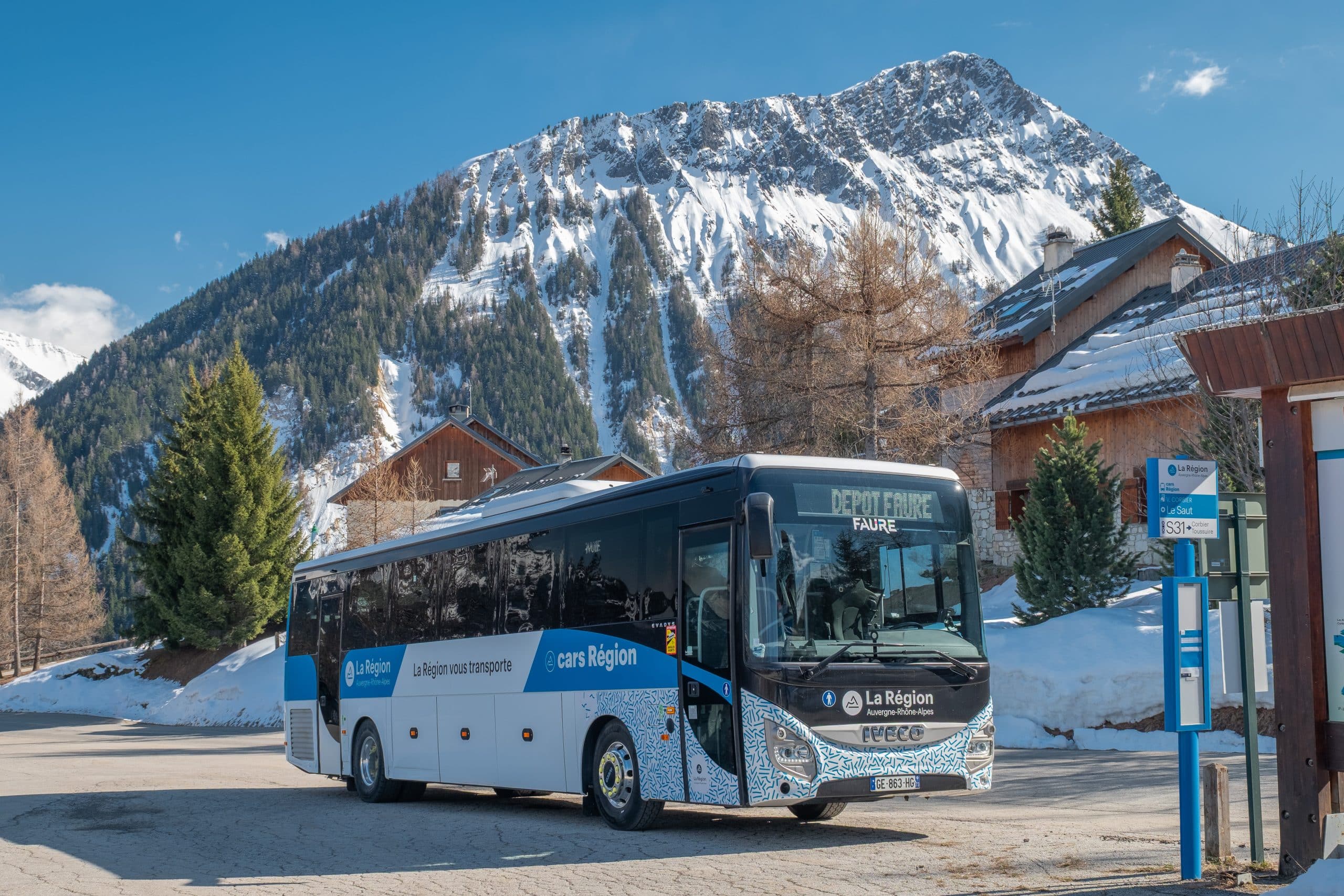 Un bus blanc et bleu est garé près d'un arrêt, dans un paysage montagnard enneigé avec chalets et forêts, sous un ciel clair. Sur l'afficheur du bus : "DEPOT FAURE". Sur le bus : "La Région Auvergne-Rhône-Alpes", "La Région vous transporte", "cars Région".