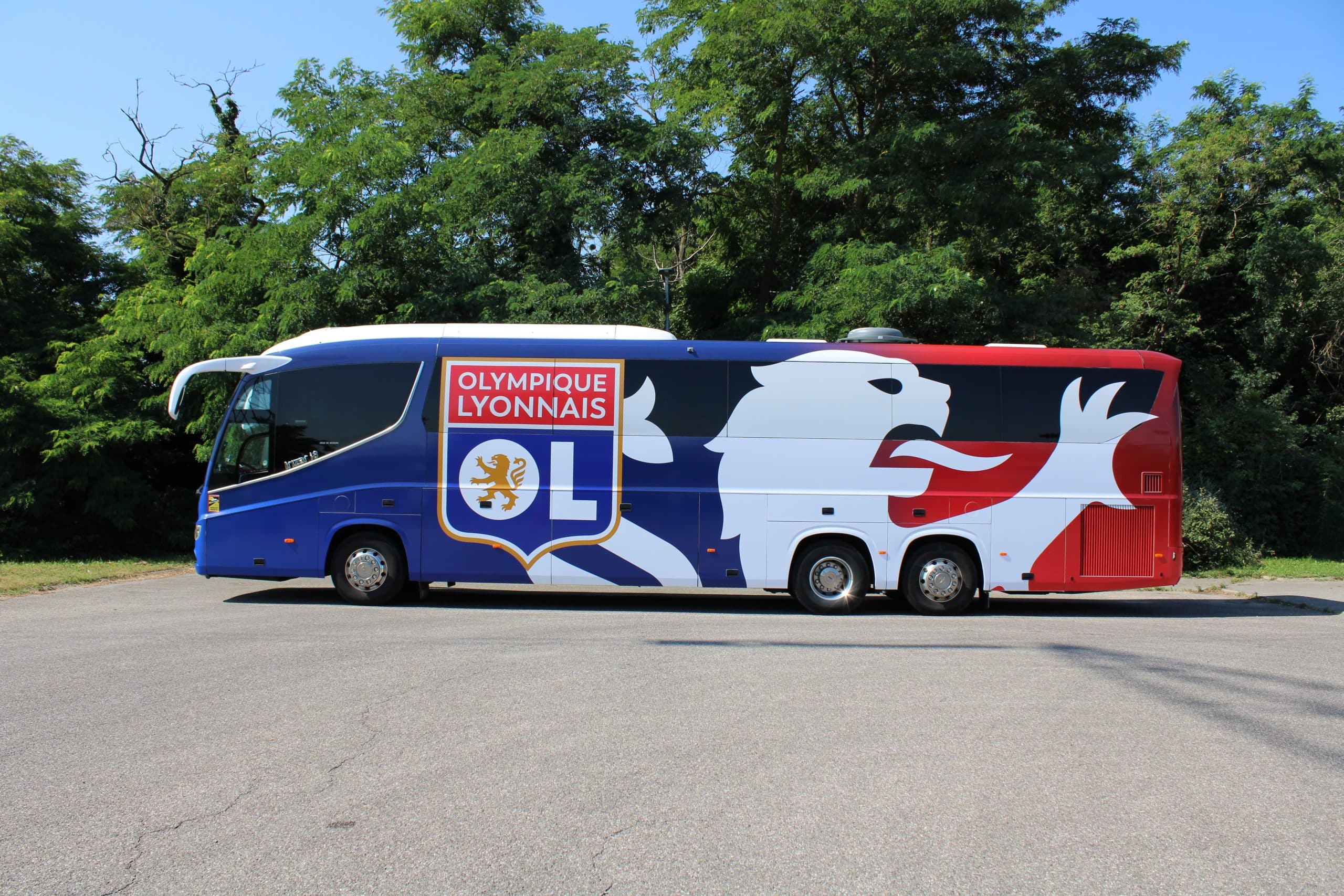 Un autocar aux couleurs bleu, blanc, rouge de l'Olympique Lyonnais est stationné sur un parking asphalté, devant des arbres verts et un ciel clair. La carrosserie affiche : OLYMPIQUE LYONNAIS, le logo OL avec un lion doré.