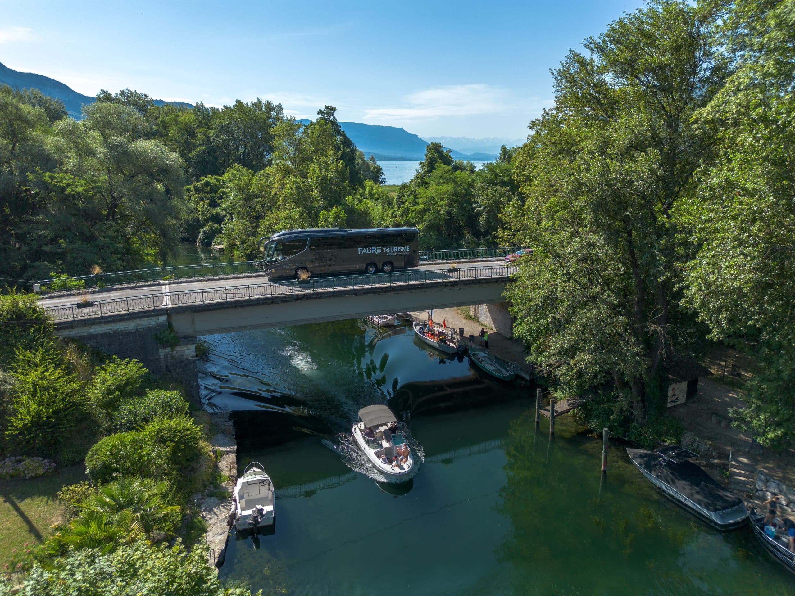 Un autocar gris FAURE TOURISME (www.faure-tourisme.fr) traverse un pont. Sous celui-ci, un bateau navigue sur une rivière bordée d'arbres, menant à un lac avec des montagnes.