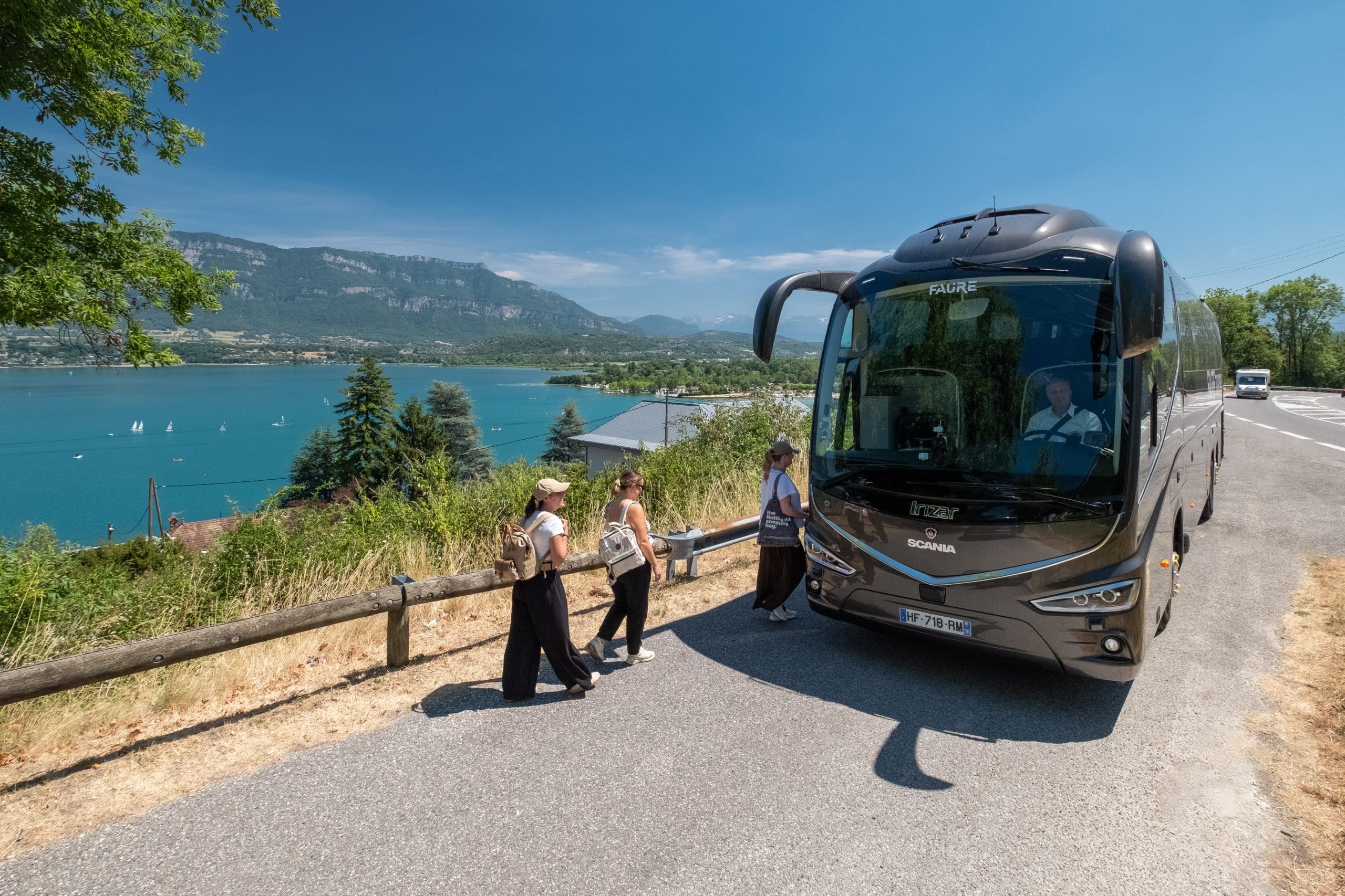 Un car de tourisme FAURE est garé sur une route, trois femmes marchent près de l'entrée. Le lac turquoise et des montagnes s'étendent à l'arrière-plan ensoleillé.