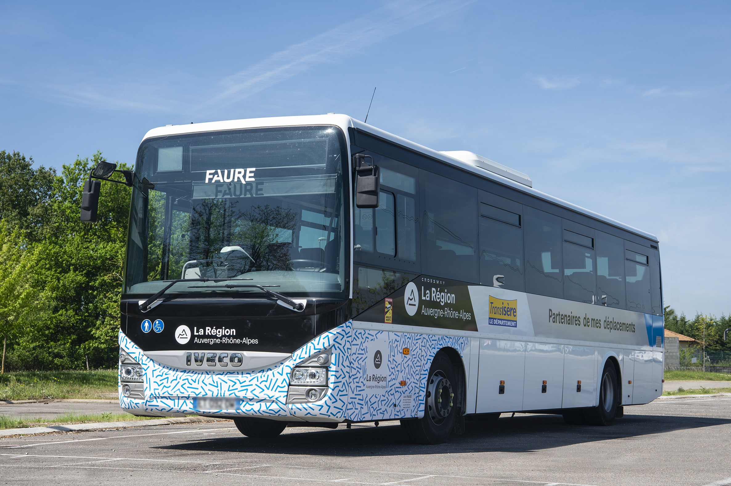 Un autocar blanc et bleu, avec des motifs géométriques bleus, est stationné sur un parking asphalté, devant des arbres verts sous un ciel bleu clair. Texte intégral : Devant : "FAURE FAURE", symboles PMR, "La Région Auvergne-Rhône-Alpes". Côté : "La Région Auvergne-Rhône-Alpes", "Transisère LE DÉPARTEMENT", "Partenaires de mes déplacements". Détail bas avant : "DANS L'ISÈRE", "SARL FAURE TRANSPORTS", "WWW.FAURE-TRANSPORTS.FR".
