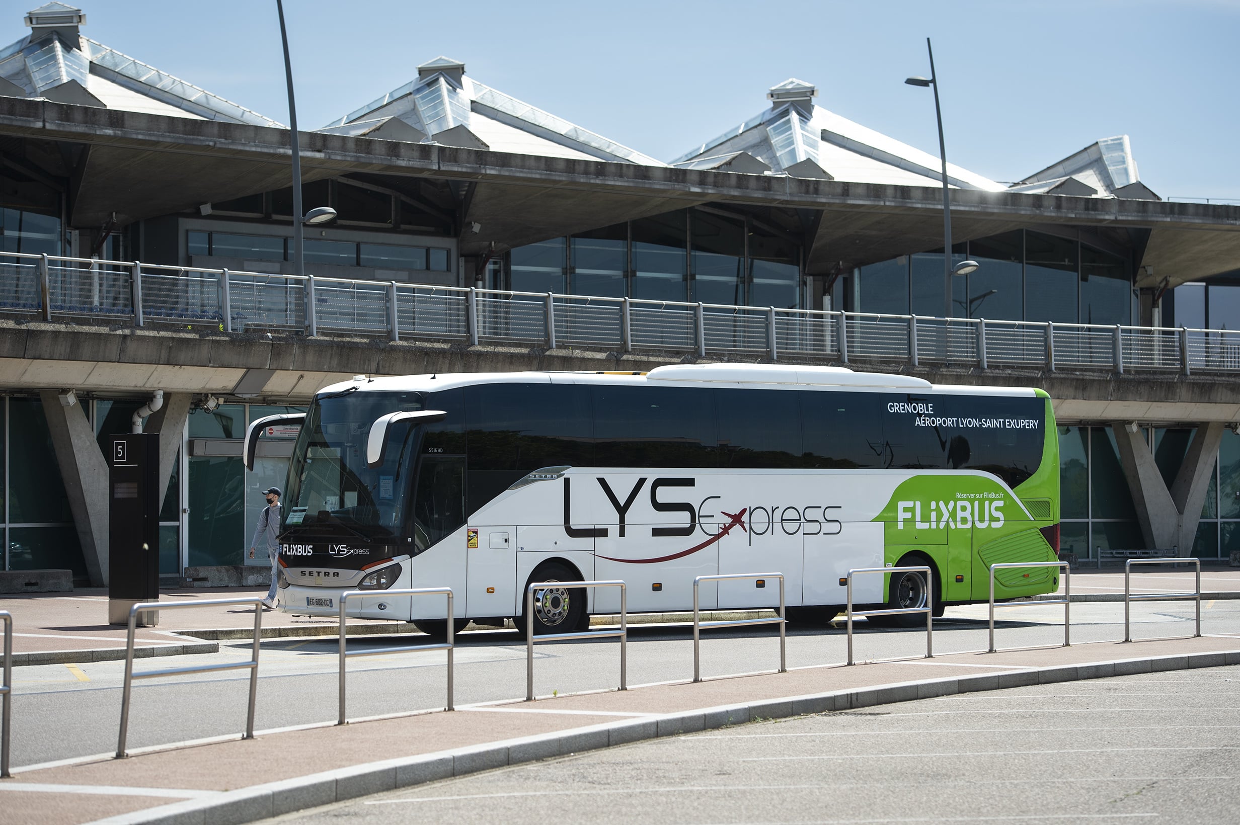 Un bus LYS express/Flixbus blanc et vert est stationné devant le terminal d'un aéroport. Texte sur l'image : FLIXBUS, LYS express, GRENOBLE AÉROPORT LYON-SAINT EXUPERY, Réserver sur FlixBus.fr.