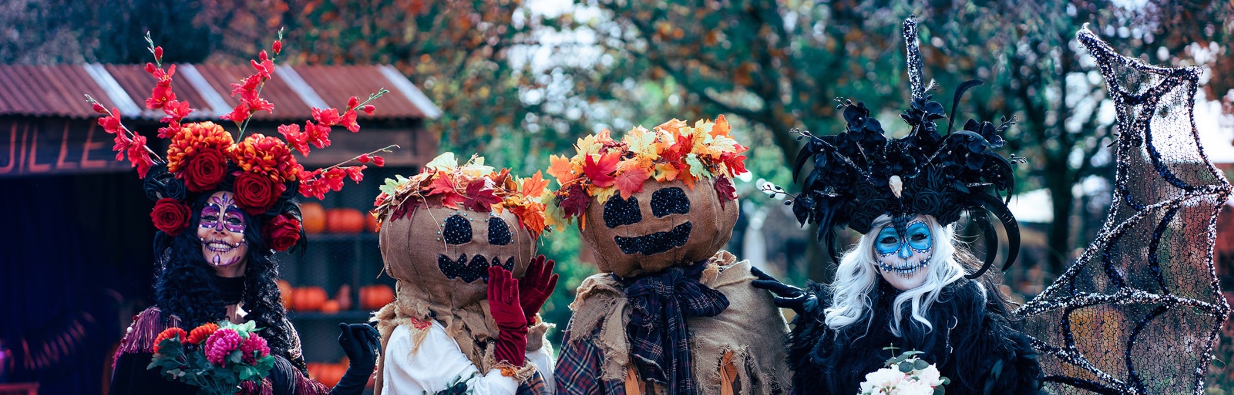 Two characters with Calavera makeup and two pumpkin scarecrows, decorated with autumn flowers and leaves, are lined up in an outdoor autumn setting.